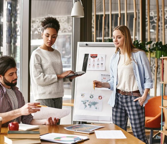 Attractive young woman standing near whiteboard with a laptop in her hands and looking to her male colleague, that holds a chart with the results. Another colleague looks at it with a smile. Concept of success